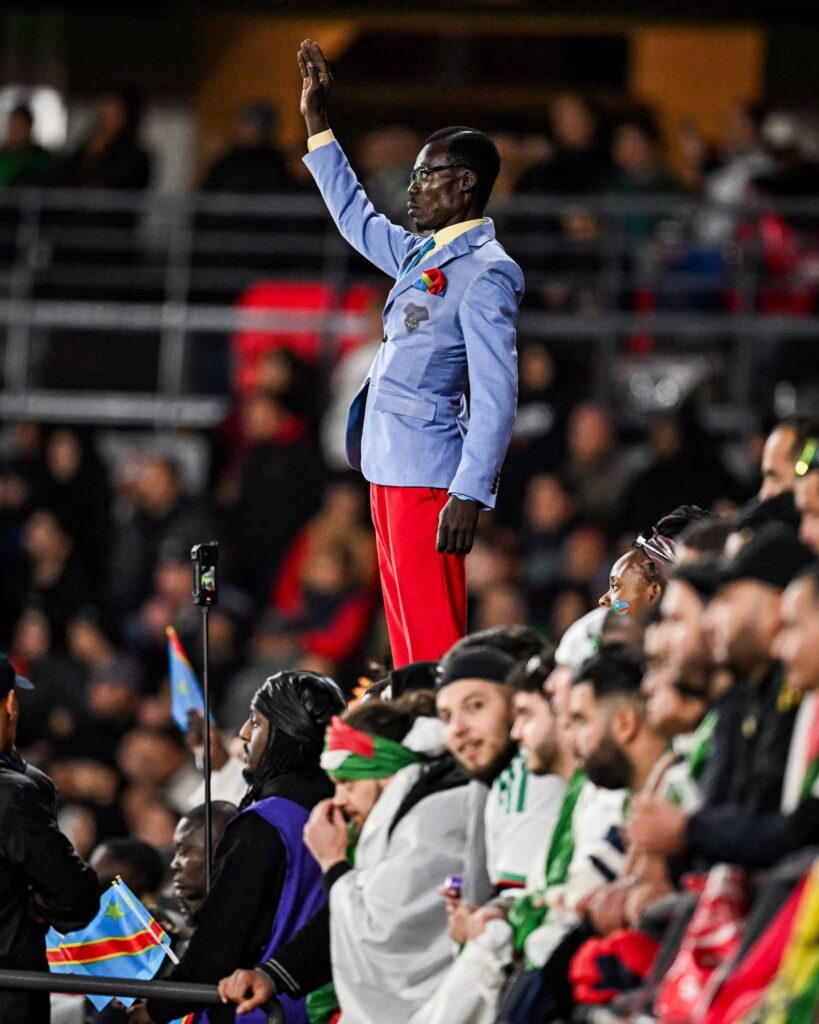 Michel Nkuka Mboladinga, also known as “Lumumba Vea,” standing motionless on a pedestal with his right arm raised, mimicking Patrice Lumumba’s statue during a DR Congo match at AFCON 2025